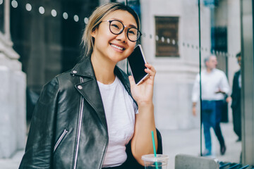 Smiling asian girl in spectacles and cool wear talking on phone sitting on bus stop bench outdoors...