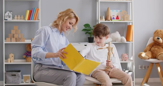 Friendly professional woman child psychologist or speech therapist sitting with little boy doing exercise in book together during therapy session in office. Psychology and education concept. 4k video