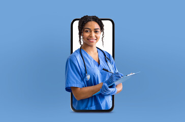 A cheerful black woman nurse dressed in blue scrubs writes on a clipboard while appearing to emerge...