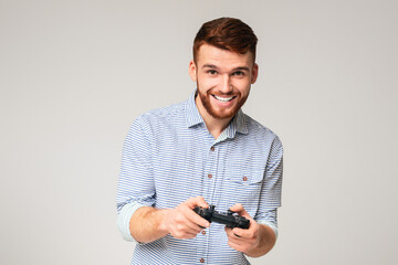 Excited millennial guy playing video games with joystick and smiling, isolated on grey studio...
