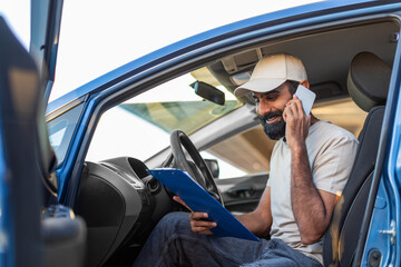 Indian man sits in the drivers seat of a blue car, engaged in conversation on his cell phone while holding a blue clipboard. He appears focused and engaged, with a relaxed demeanor
