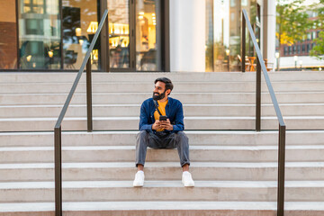 A young Indian man is sitting on the steps of a modern outdoor staircase in an urban area. He...