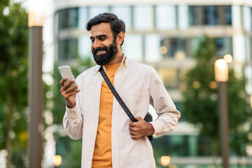 Indian man stands outdoors in an urban park smiling as he looks down at his smartphone. He is...