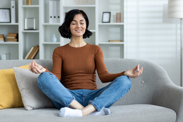 Smiling young woman relaxing on sofa at home, relaxing with yoga, sitting in lotus position with...