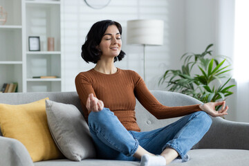 Fototapeta premium Young girl in lotus pose doing yoga, sitting on sofa at home with closed eyes and meditating
