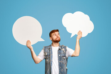 Young redhaired man having complex choice holding two speech bubbles on blue background, mockup, empty space