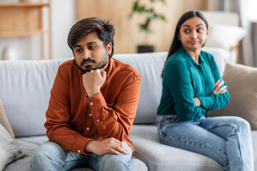 A distressed Indian couple sits on a sofa; the man looks contemplative and sad while the woman...