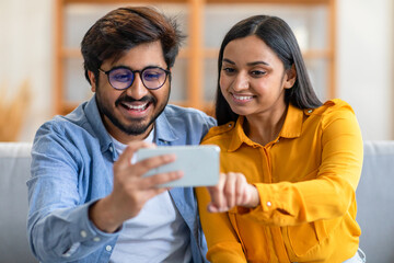 A cheerful Indian couple sits on their couch, smiling warmly as they take a selfie together. They are enjoying a relaxed moment indoors.