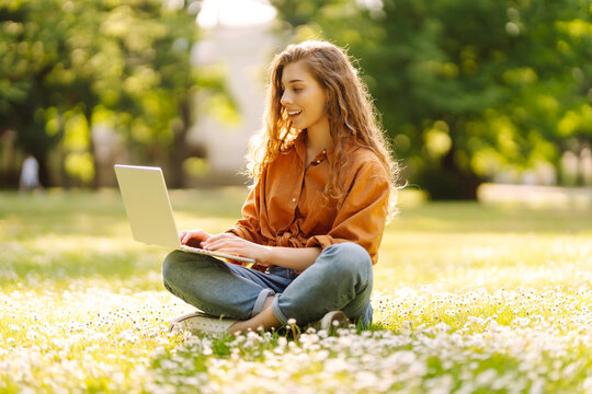 Young woman with laptop sits on the grass in the park on a sunny day.  Online training, remote work. Education online.