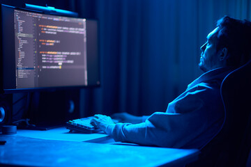 A coder is working late at night on a computer screen, coding and using a smartphone app