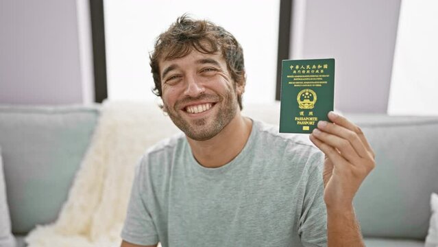 Cheerful young man, casually sitting on the living room sofa, joyfully holds his macao passport indoor, displaying a confidence-filled, happy smile at home.