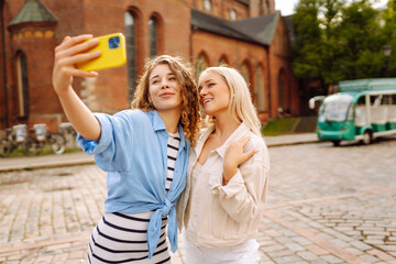 Fototapeta premium Two young smiling women in summer clothes taking selfie self portrait photos on smartphone. Models posing in the street. Fashion, travel, technology, blogging concept.