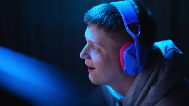 Closeup face of happy excited cyber player wearing gaming headset having fun playing online video games on personal computer in dark room with blue neon light. Concept of modern online entertainment.