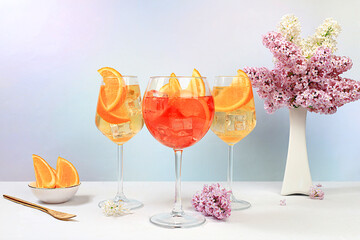 Festive alcoholic cocktail Aperol spritz in glasses on a bright background with a bouquet of lilacs, summer bar concept, alcoholic drinks at a party, restaurant and cafe advertising,
