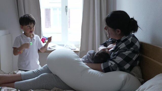 Mother breastfeeding newborn while older child looks on, illustrating the dynamics of family life, sibling curiosity, and the nurturing role of a mother, emphasizing the importance of maternal care
