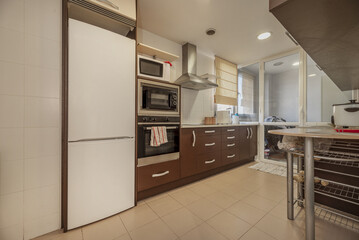 A kitchen with brown wooden furniture with a glazed terrace in the background and a column with integrated appliances