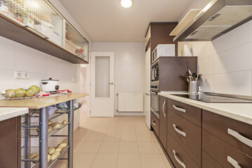 A kitchen with brown wooden furniture, a column with integrated appliances, a light countertop and...