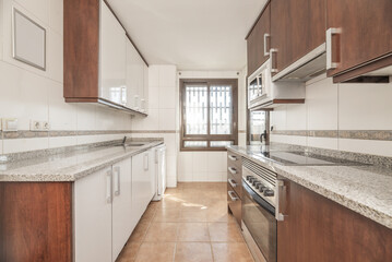 A kitchen with combined brown and white wooden furniture, with integrated appliances, gray granite countertops and brown aluminum windows and light brown stoneware floors