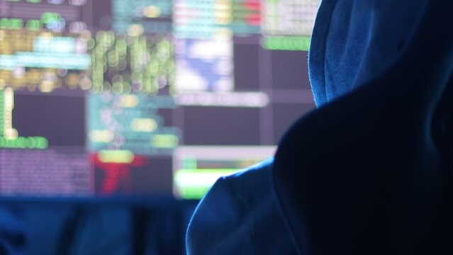 In a dimly lit room, a cloaked figure types code on a computer, hinting at a mysterious hacker