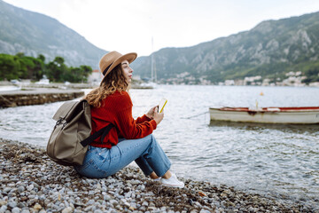 Young female traveler with backpack and in warm wearing standing on quay against lake and looking at phone screen. Travelling, lifestyle, adventure, concept.