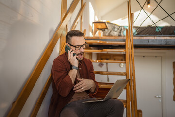 Adult man sit and talk on mobile phone and work on laptop on stairs