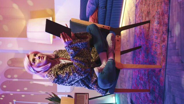 Portrait of stylish girl working at home during vacation. Disco neon stroboscopes illuminate room. Beautiful fashionable glamorous young woman sitting on chair using modern digital tablet for work.