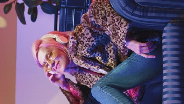 Beautiful young creative girl in wearless headphones listening to music, dancing, resting on soft vintage sofa. Trandy cool woman in sunglasses posing in front of camera, smiling in retro studio.