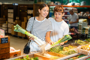 Enthusiastic young vegetarian couple standing in vegetable section of local grocery store,...