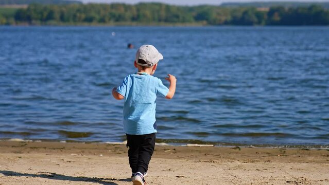 Rear view of a toddler boy running towards the river. Kid carries a stone and throws it into water.