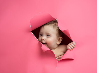 Cute Caucasian newborn baby boy peeks out of a hole in a paper pink background.