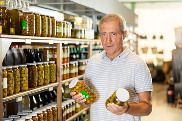 Mature man in supermarket selects examines compares two cans jars of olives. Male customer chooses...