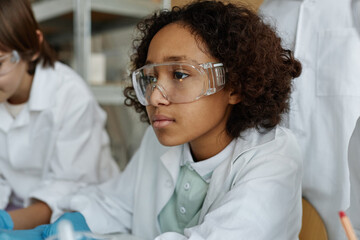 Medium closeup portrait of African American boy wearing lab coat and goggles learning Chemistry at...