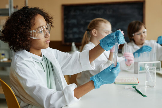 African American schoolboy wearing lab coat sitting at table in classroom with classmates doing laboratory experiment during Chemistry class