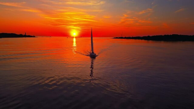 Aerial view of a sailboat peacefully sailing at sunset on calm waters under an orange sky, creating a serene setting