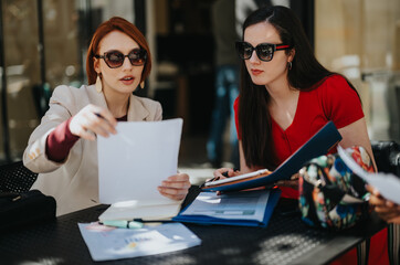 Two focused businesswomen reviewing documents and strategizing at an outdoor meeting space, collaborating on project ideas.