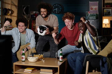 Multi-ethnic group of excited young men spending evening together in living room watching soccer...