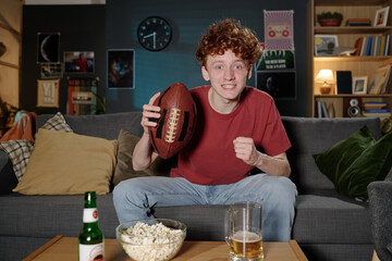 POV of cheerful red-haired Caucasian guy holding ball sitting on sofa in living room watching...