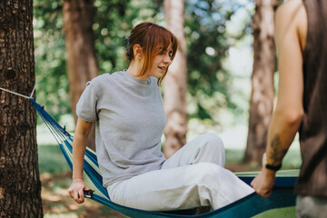 A young woman smiles while sitting in a hammock, interacting with a friend in a lush park on a...