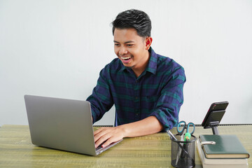 cheerful young asian man in blue shirt sit work at wooden desk with pc laptop isolated on white...