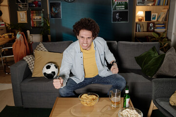 High angle view of young biracial soccer fan sitting on couch in living room watching match on TV...