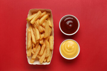 Paper box with tasty french fries and bowls of sauce on red background