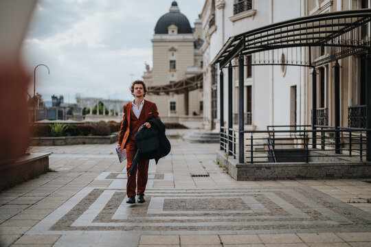Confident businessman in orange suit walking outside modern building carrying a briefcase on a cloudy day. Professional attire, city background.