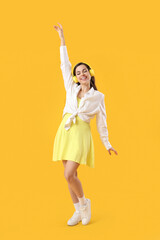 Pretty young woman in modern headphones listening to music and dancing on yellow background
