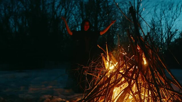 Witch performing witchcraft at bonfire. Mysterious female conjuring in forest during wintery night. She is doing rituals on snow covered landscape on halloween.