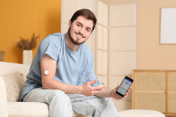 Diabetic young man with glucose sensor, glucometer and mobile phone sitting on sofa at home