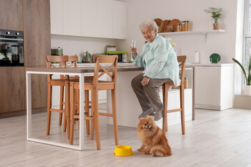 Senior woman with glass of water and Pomeranian dog in kitchen