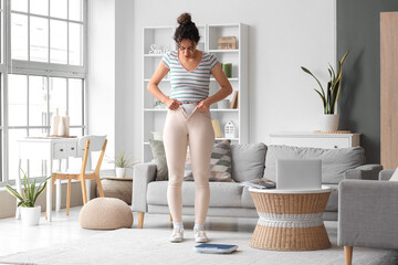 Young African-American woman trying to button tight pants at home. Diet concept