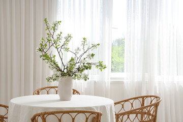 Vase with blooming branches on table near window in dining room