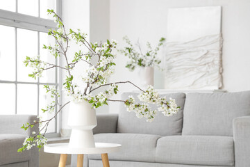 Vase with blooming branches on chest of drawers near grey sofa in light living room