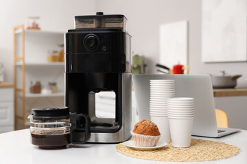 Modern coffee machine, muffin, paper cups and laptop on white table in kitchen